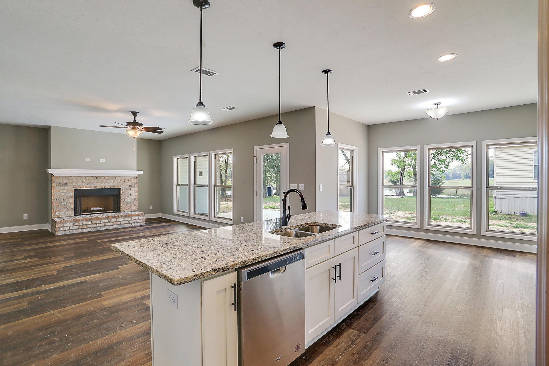 Open-concept kitchen and living room featuring a brick fireplace with metal basket, granite countertops, stainless steel dishwasher with black handle, white cabinetry, and hardwood