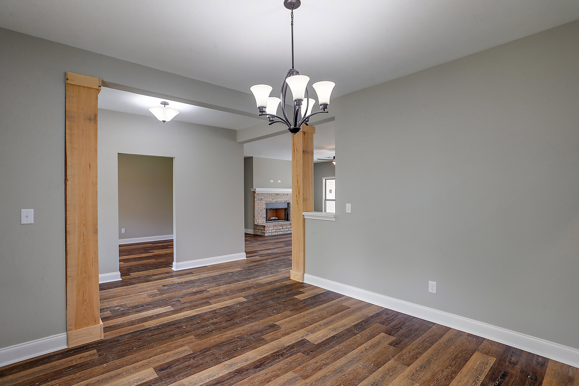 Open living room with wide plank wood flooring, ornate chandelier with white lamps, plaster walls, fireplace beside tall window, and open doorway.