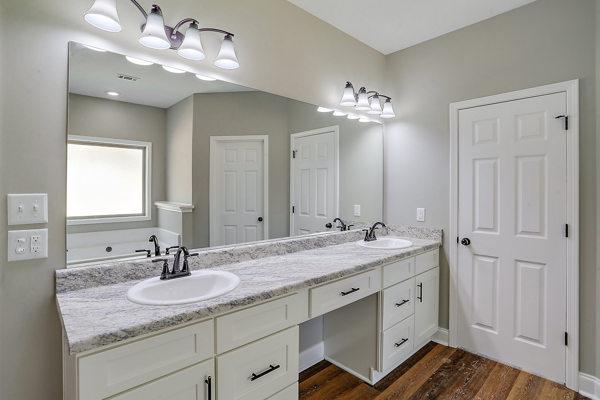Bathroom with double vanity featuring undermount sinks, quartz countertop, large framed mirror, white cabinetry with silver hardware, three-light fixture above, white door with