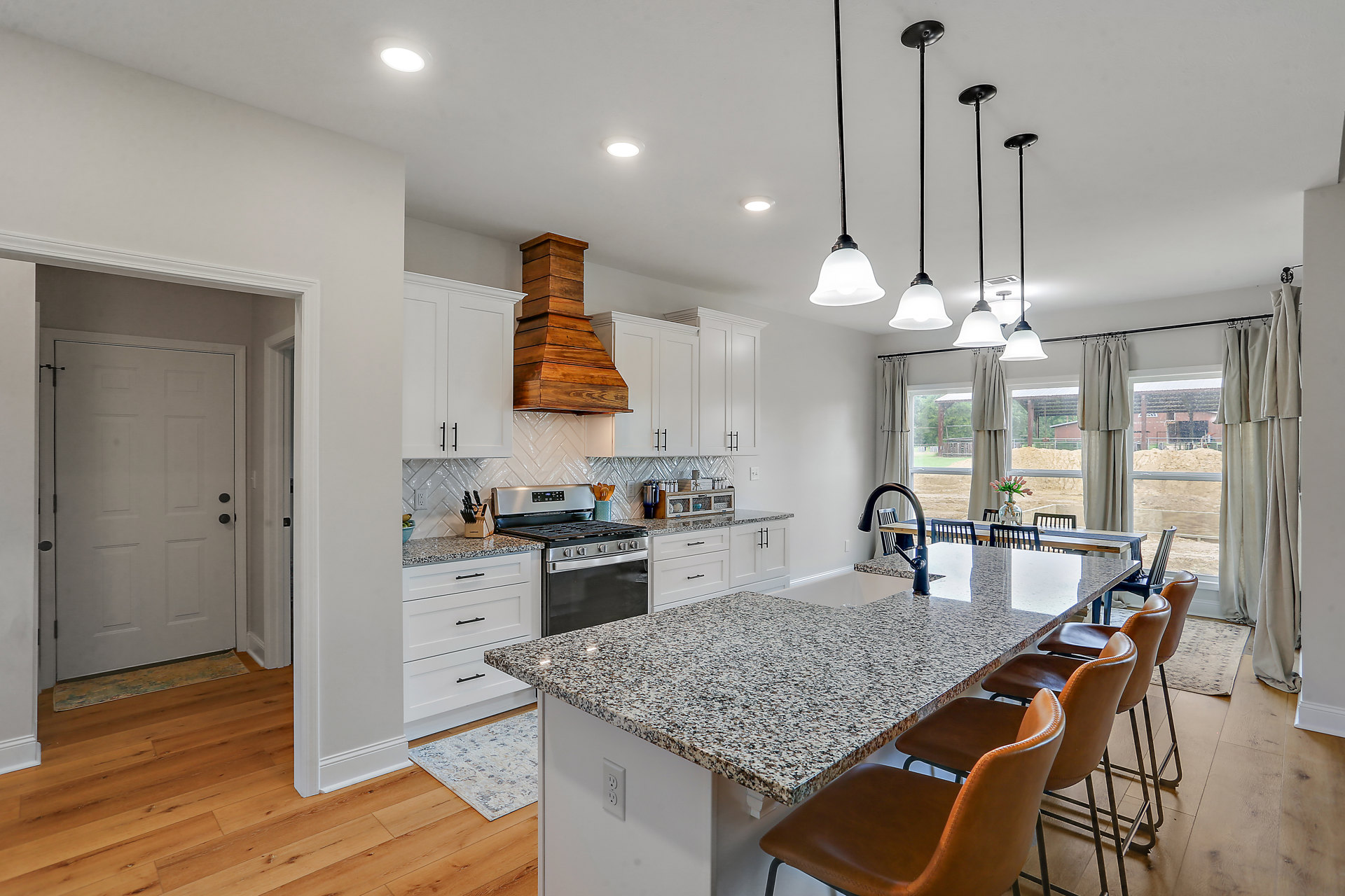 Spacious kitchen featuring a large central island with barstool chairs, light stone countertops, white cabinetry, stainless steel sink, wood range hood, vase of flowers, and a