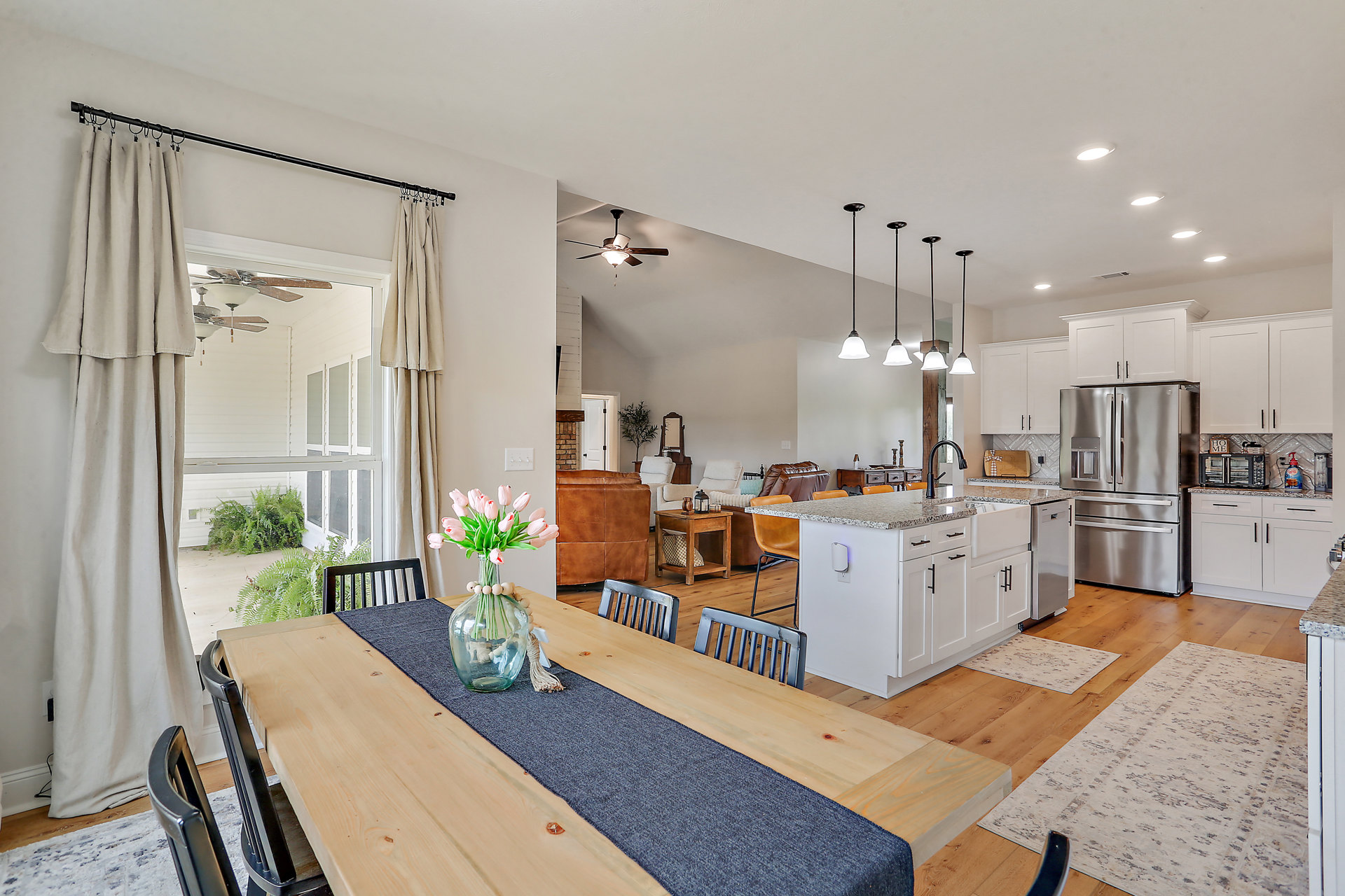Dining area with wooden table, glass jar of pink tulips, open doorway leading to kitchen featuring white cabinetry, stainless steel refrigerator, and light countertops, sheer white
