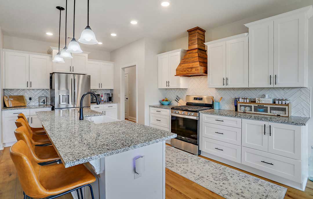 Marble kitchen countertop with stainless steel double-door refrigerator, stove, wood chimney, black lamp on counter, chairs tucked under dining table, white cabinetry and drawers