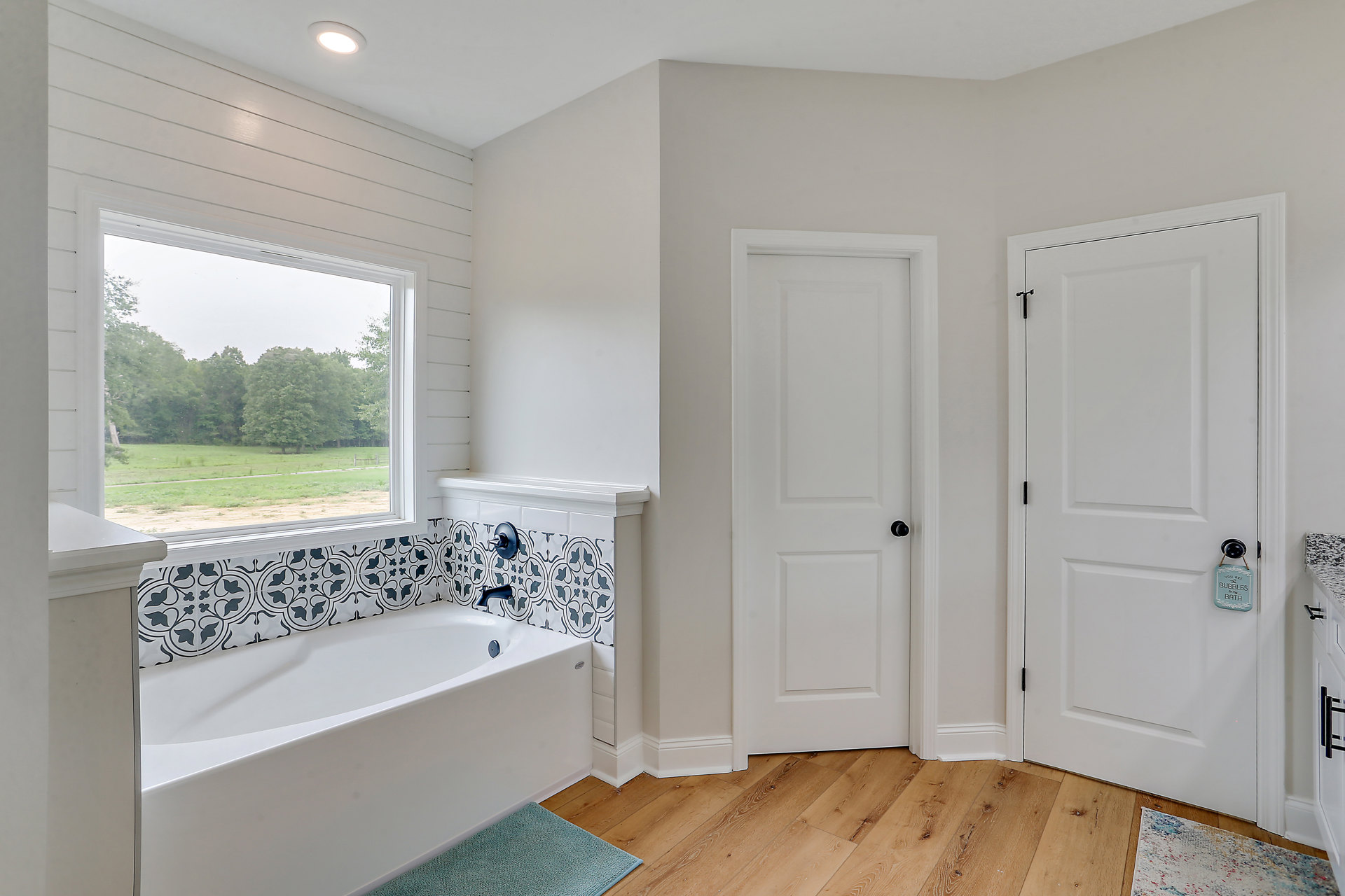 Bathroom with freestanding bathtub, white door with black knob and sign, second door, window showing trees, light-colored flooring, neutral walls