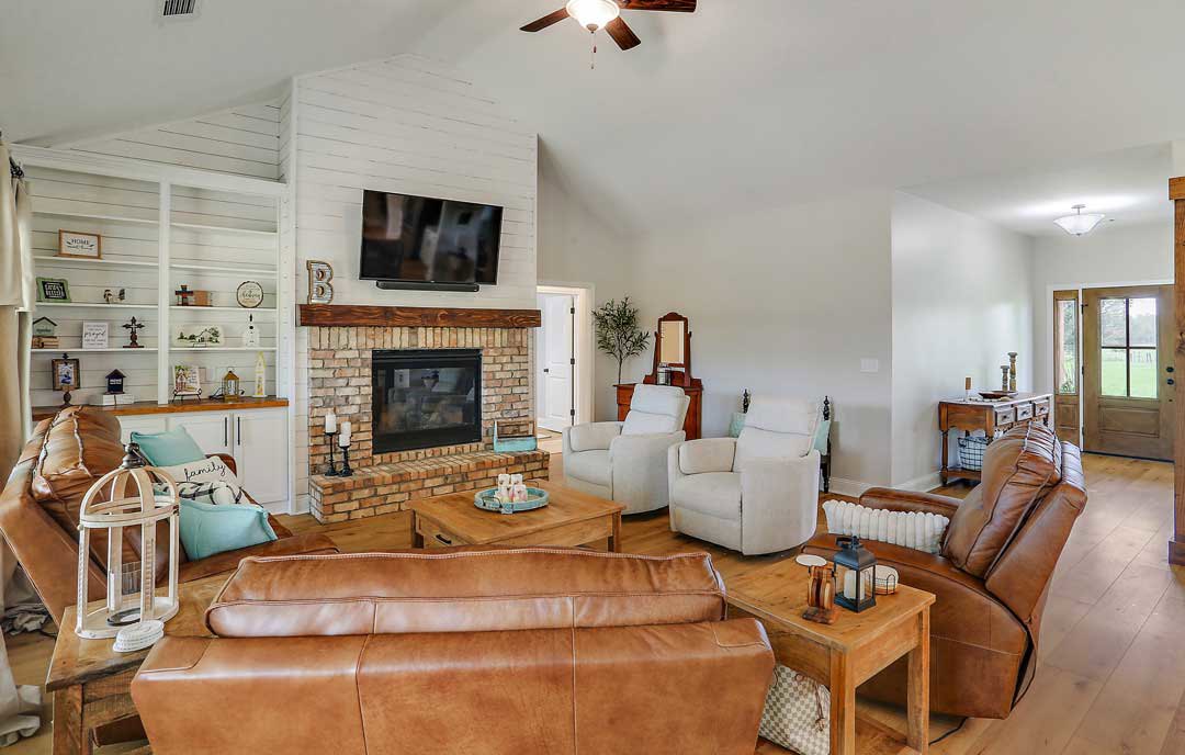 Brown leather couch and white chair arranged on wood flooring in living room with brick accent wall, fireplace, television on table, and large window.