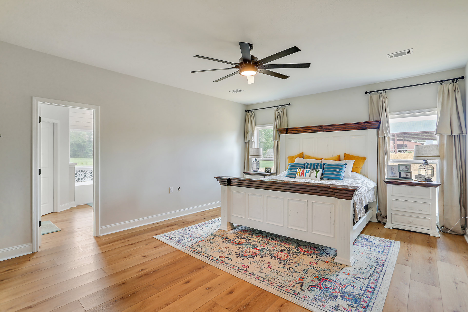 Bedroom with a large upholstered bed, ceiling fan with light, white curtains, bedside lamp, white dresser topped with picture frame, wood laminate flooring, area rug near door