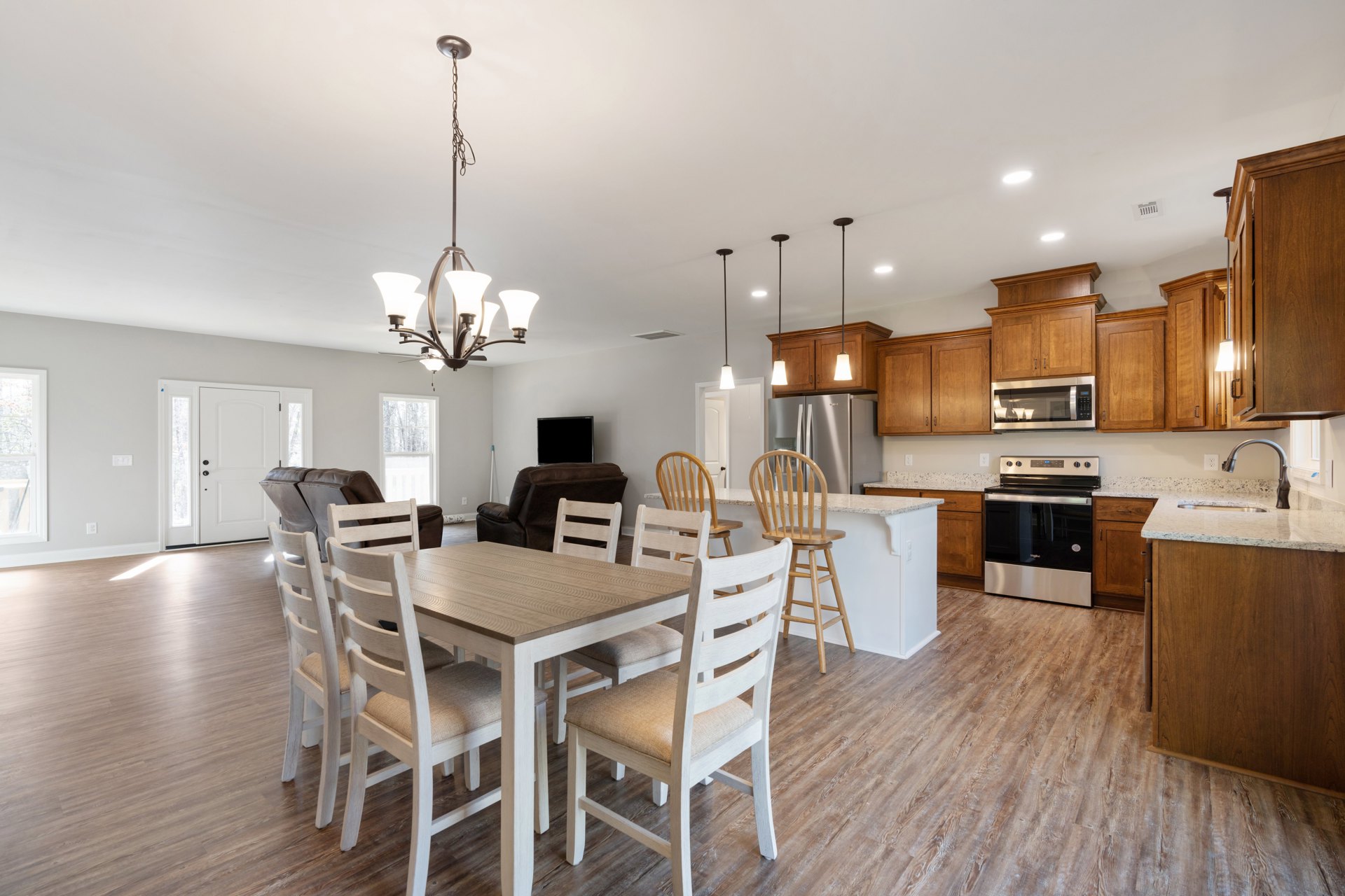 Dining area with white cushioned chairs around a wooden table, adjacent to a modern kitchen featuring black glass stove, white cabinetry, stone countertops, and built-in microwave.