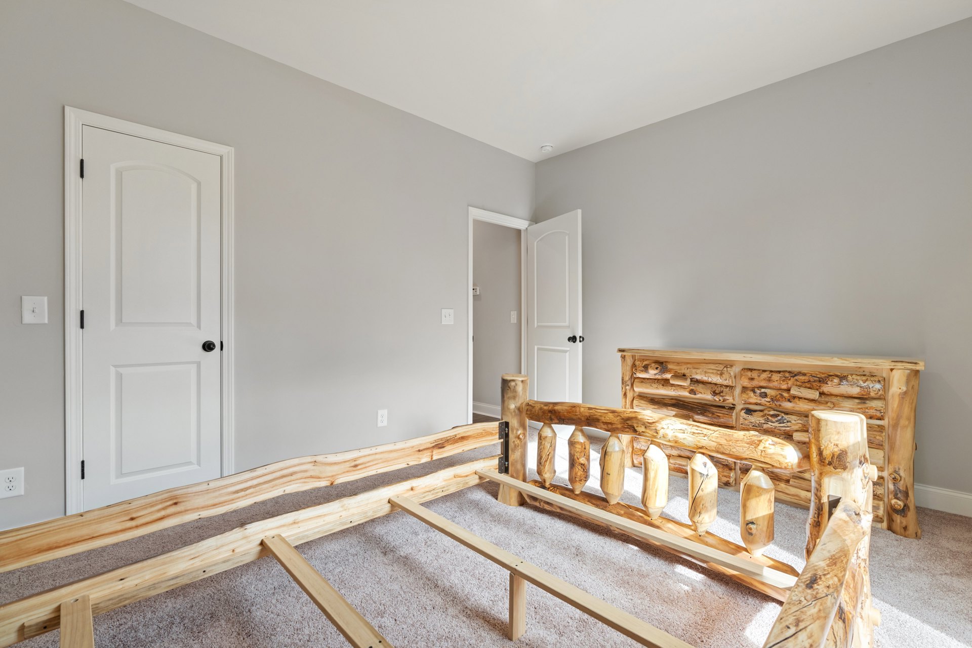 Wooden bed frame on carpeted floor beside white door with black knob and light switch on plaster wall