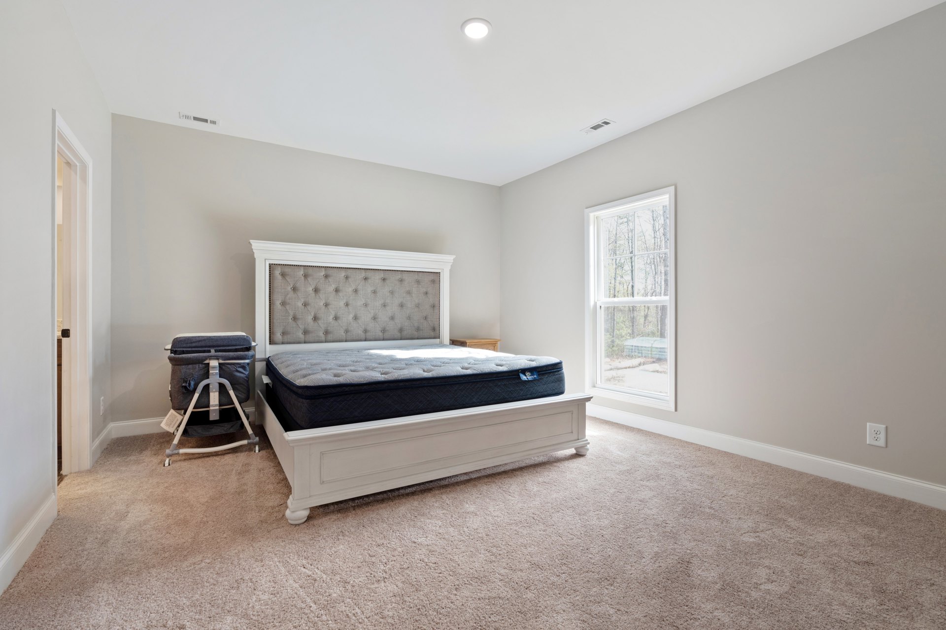 White baby crib with cloth cover and mattress on carpeted bedroom floor, headboard close up, window with trees visible outside, plaster walls, pillows and bedding