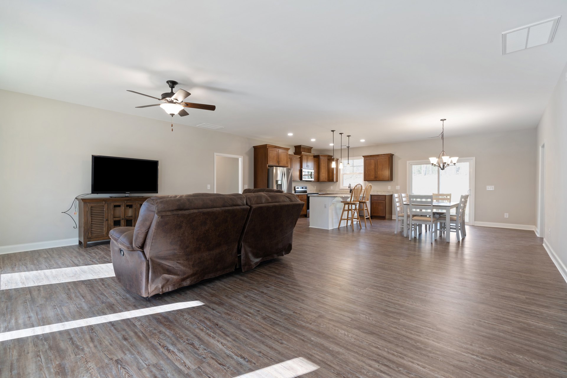 Brown leather couch with white stitched back on wood flooring, white table in front, open kitchen visible in background, white ceiling with vent and ceiling fan with light fixture