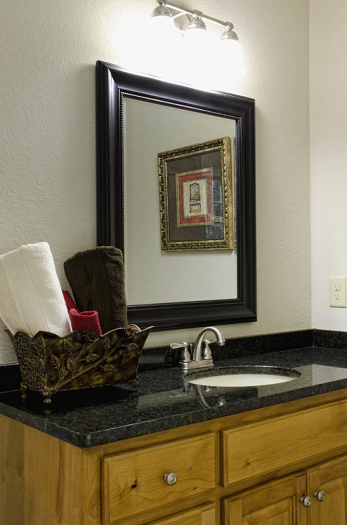 Rectangular mirror above a white sink with chrome faucet, marble countertop, metal basket holding folded towels, framed artwork on wall, and decorative object beside brown sweater.