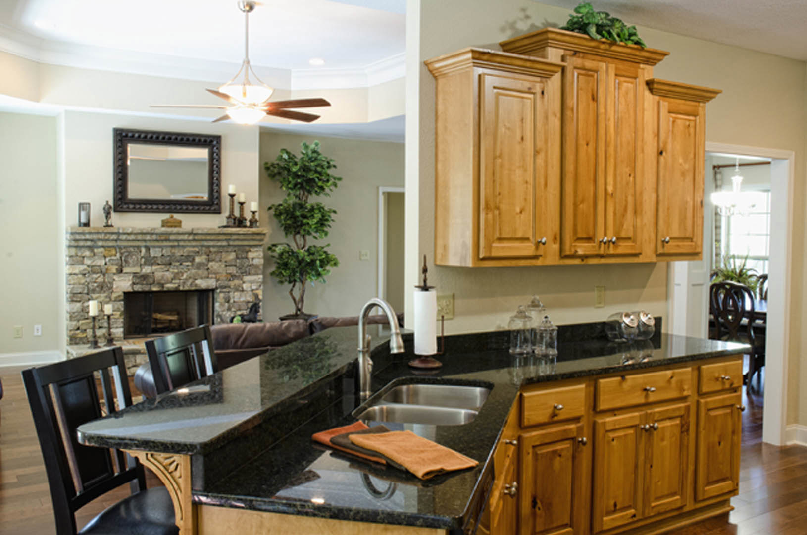 Open-concept kitchen featuring a farmhouse sink, stone fireplace, wooden cabinetry with a potted plant, dining table, ceiling fan, wall mirror, and light countertops.