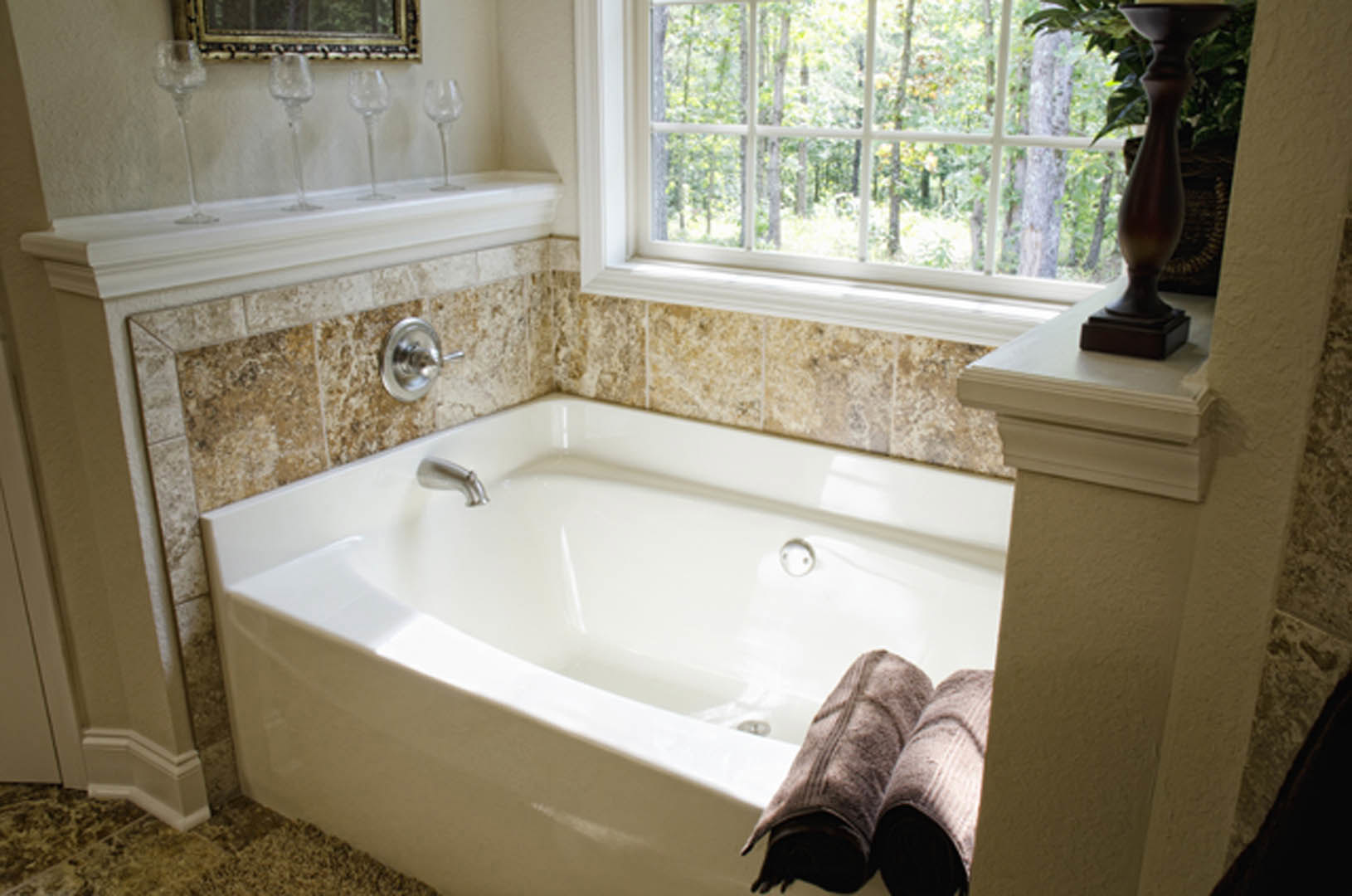 Freestanding white bathtub with chrome faucet, gray tile walls, large window, towel hanging on wall, and modern sink visible in custom bathroom