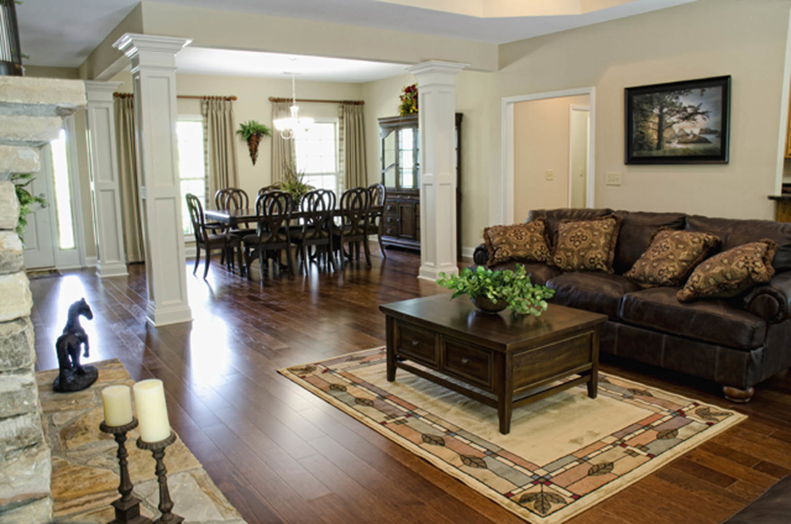 Open-plan living room with brown leather couch, accent pillows, wooden dining table, framed tree artwork on wall, coffee table with decor, hardwood floors, and neutral walls.
