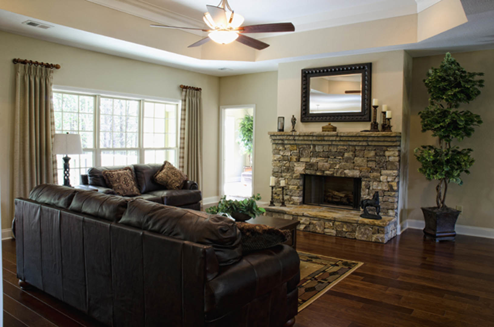 Living room with light-colored couch, stone fireplace topped with horse statue, potted plant beside hearth, wall mirror, ceiling fan, hardwood floors