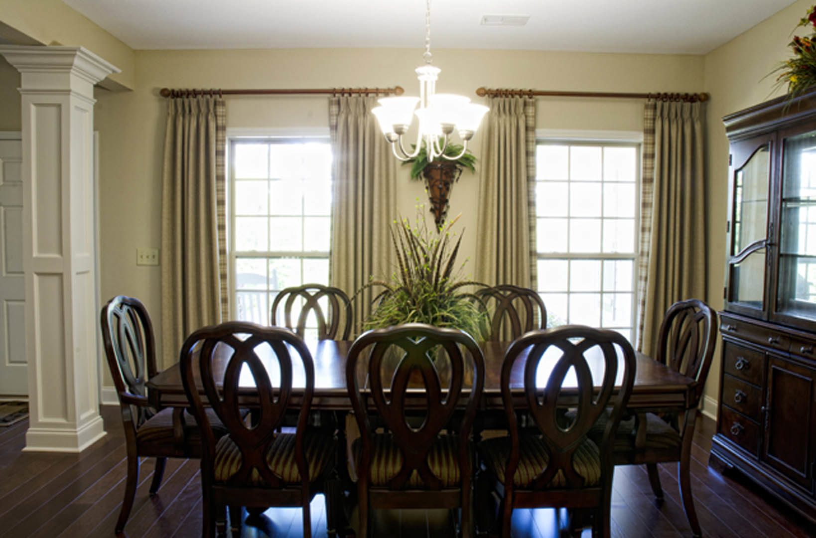 Wood dining table with upholstered chairs, potted plant centerpiece, white paneled door, large windows with sheer curtains, light hardwood floor, neutral walls, and recessed