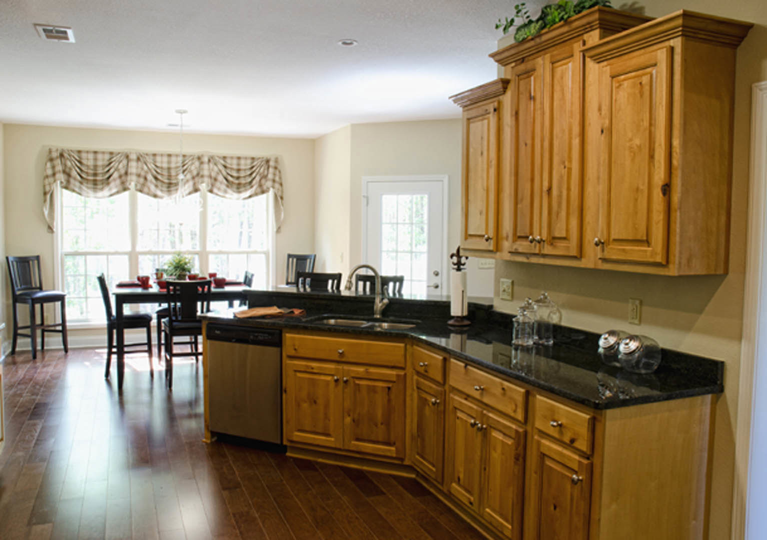 Kitchen with natural wood cabinets, white countertops, dining table with chairs, brown and white checkered curtain, hardwood flooring, and a potted plant on the table