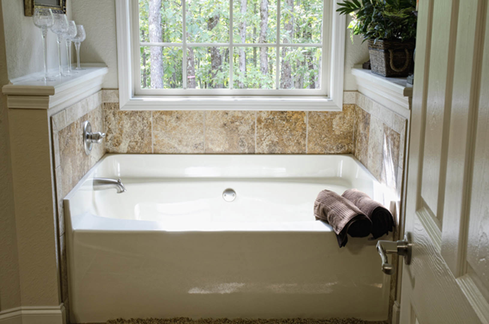 Freestanding white bathtub with a folded towel draped over the edge, potted plant in woven basket nearby, large window and neutral bathroom finishes