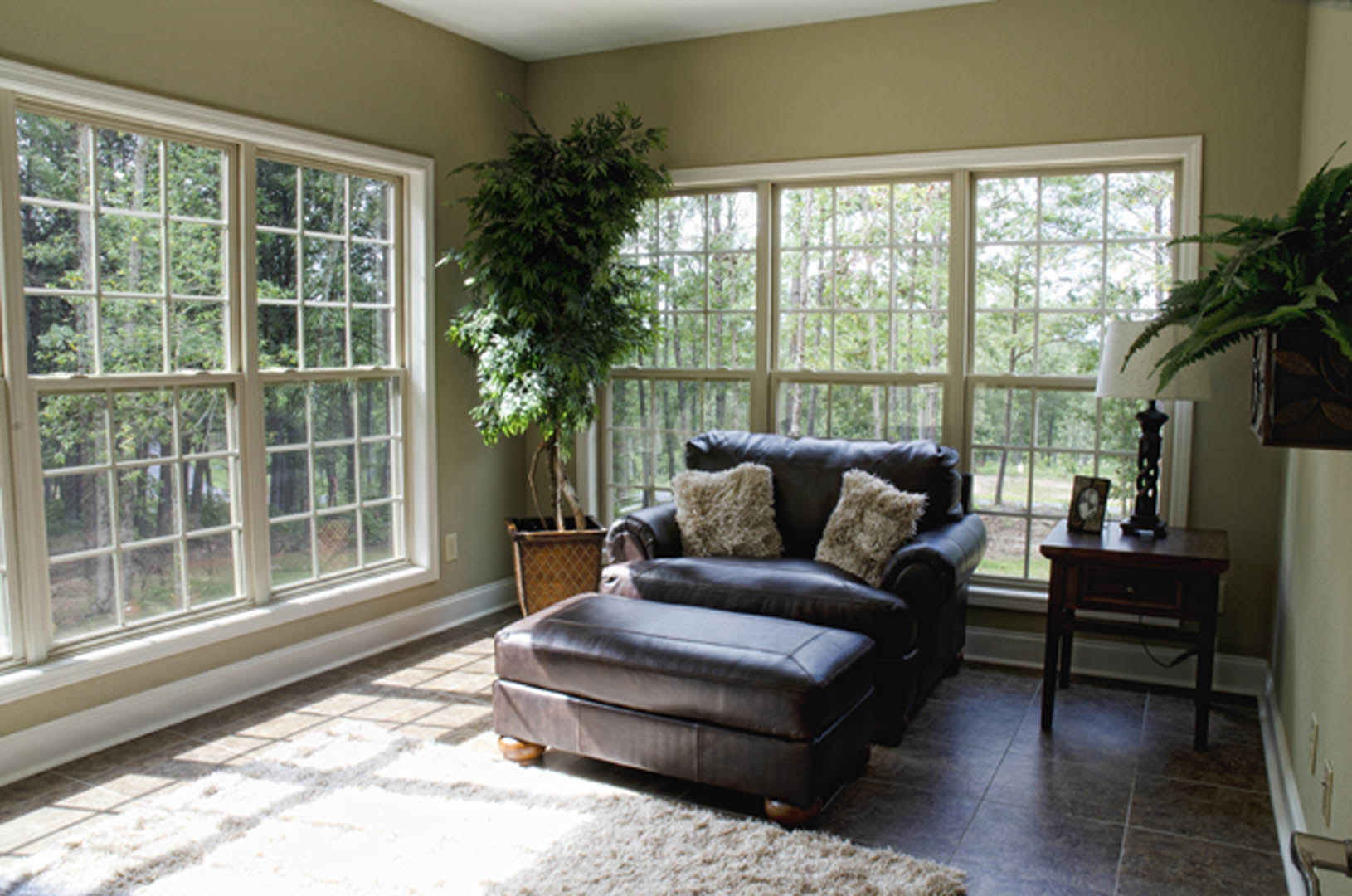 Living room with a brown leather chair and ottoman, decorative pillows, potted tree near window, wooden side table with drawer and phone, light-colored flooring and neutral walls
