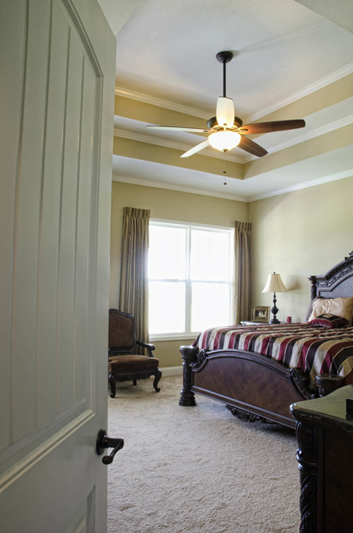 Bedroom with striped bedding, ceiling fan with light fixture, window with curtains, bedside lamp on table, upholstered chair in foreground.