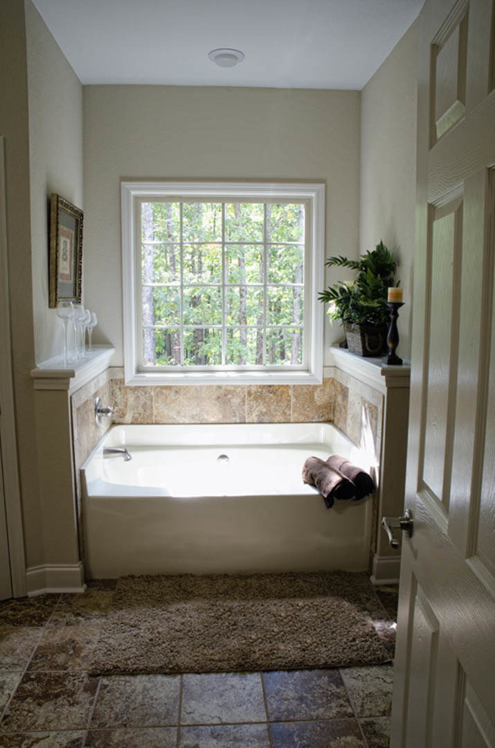 Freestanding white bathtub beside a potted plant and candle, light-filled bathroom with neutral walls and tiled floor