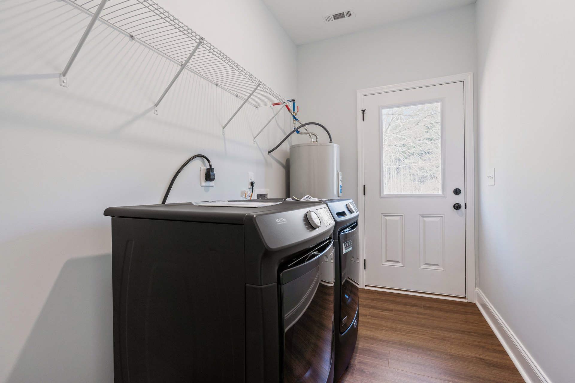 Front-loading washer and dryer set in a laundry room with white cabinetry, black oven, white door with window, and a window overlooking trees; white water tank with top valve