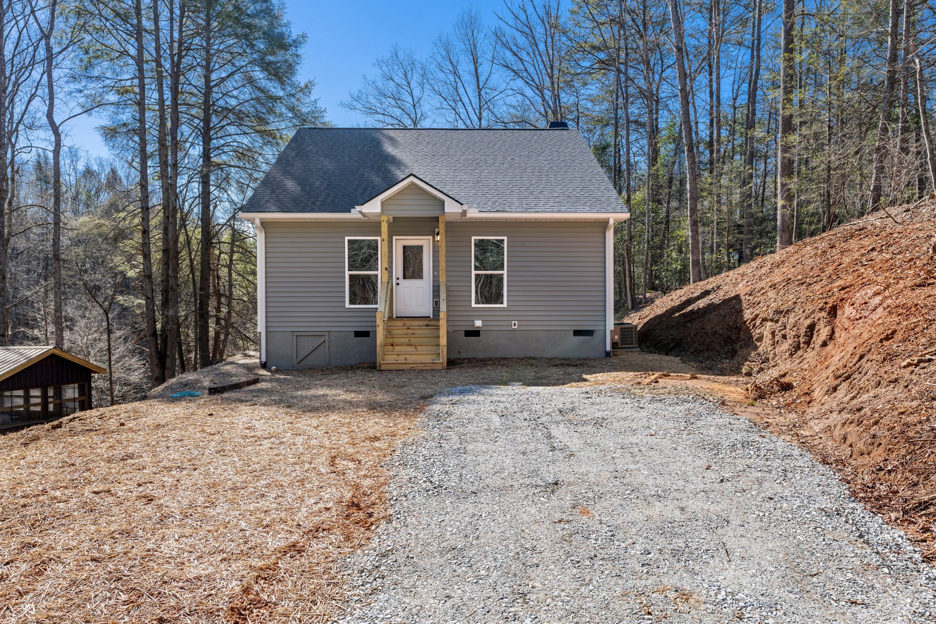 Gravel driveway leading to a cottage-style home with a white door, wooden staircase, and windows; surrounded by mature trees and greenery.