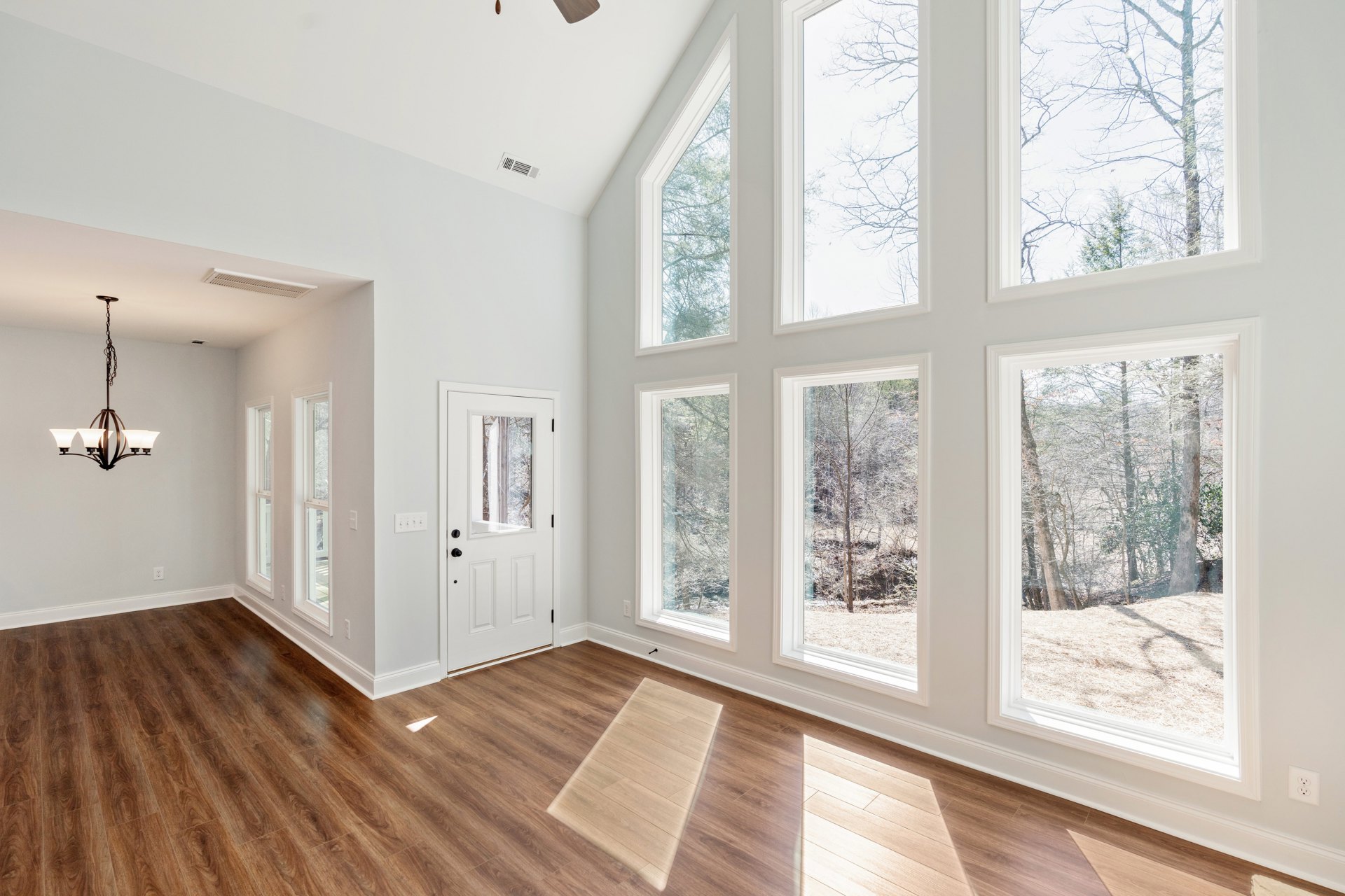 Wood flooring in a bright room with white door, multiple windows, ceiling fan, and chandelier