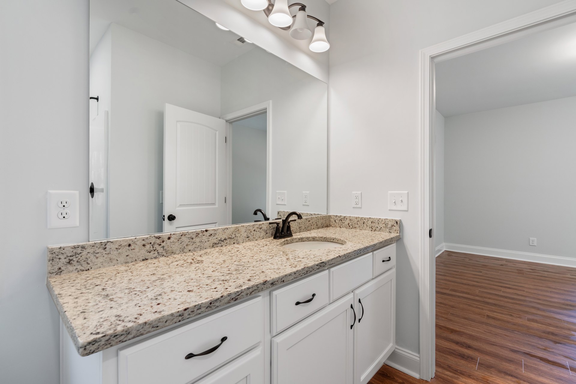 Bathroom with marble countertop, large mirror above white cabinets with black handles, wood flooring with white trim, black hardware on white door, electrical outlet visible near