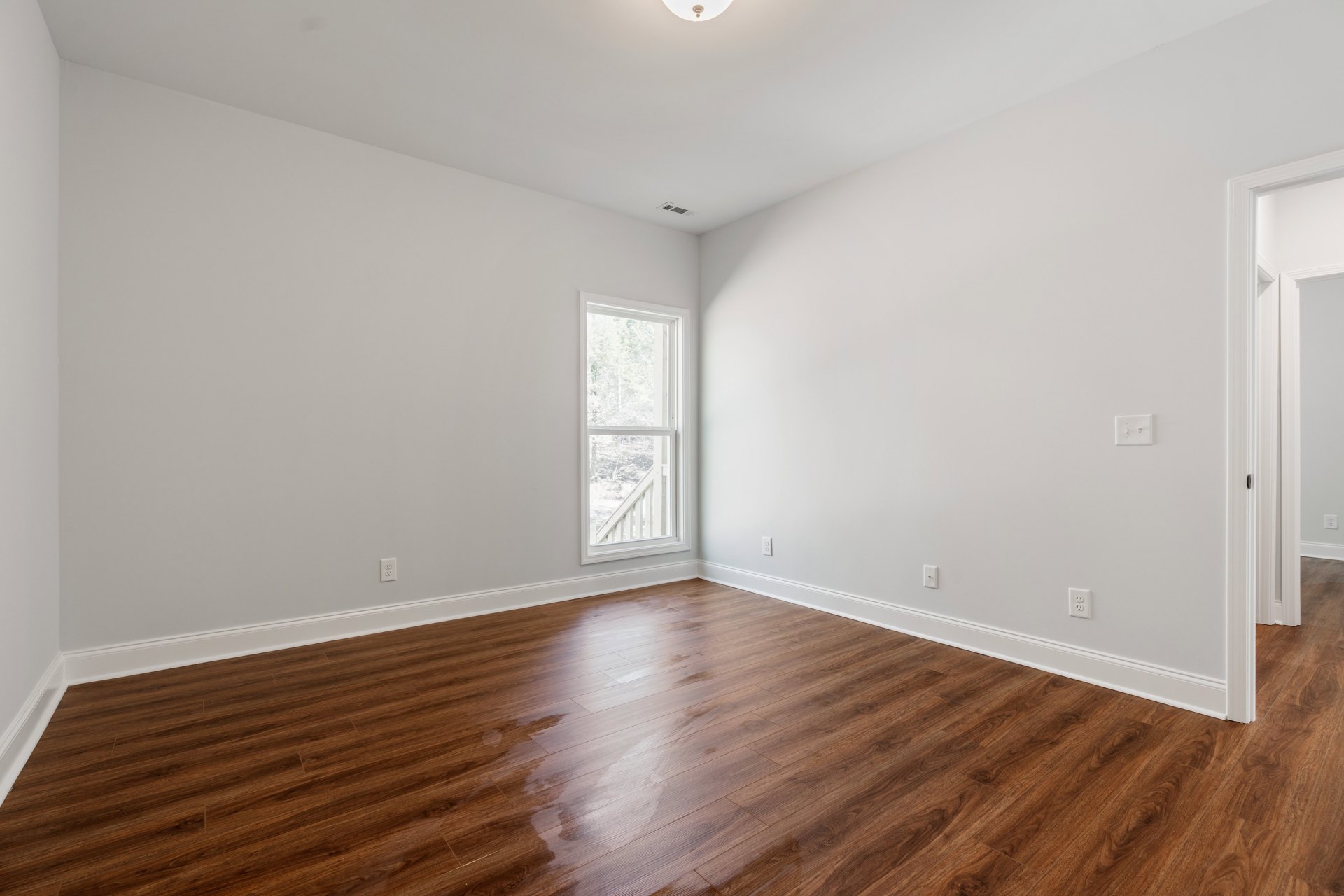 Sunlit room featuring wet hardwood flooring, plaster walls, and a large window overlooking leafy trees.