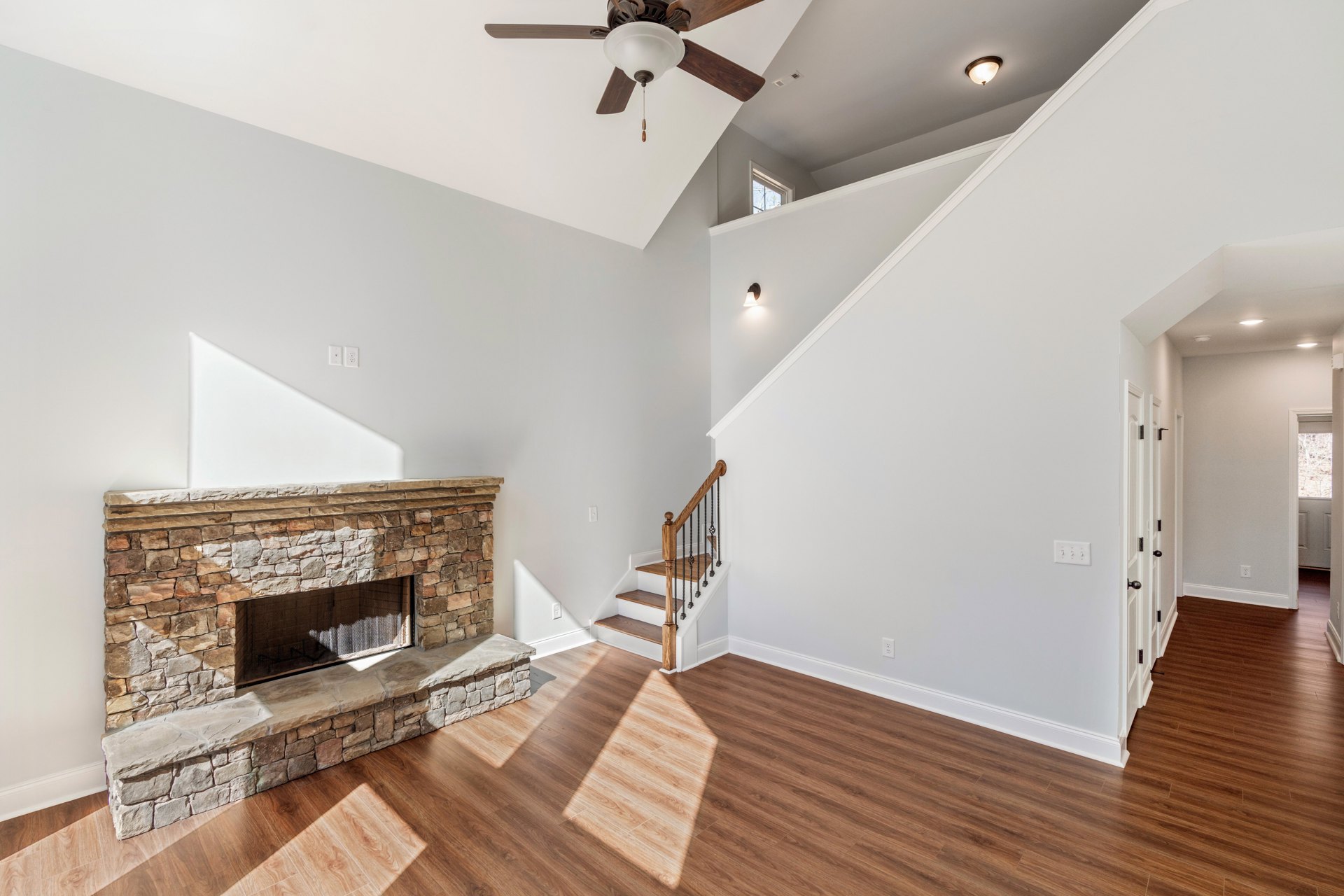 Living room with stone fireplace, wood flooring, ceiling fan, white walls, and wooden staircase with metal railings