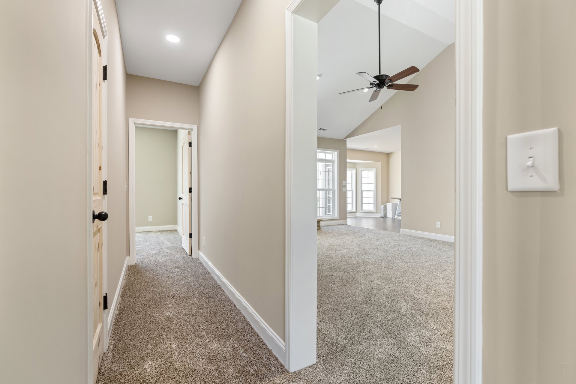 Hallway with beige carpet flooring, white walls, ceiling fan with light fixture, white door with glass panes, and a window with divided lights