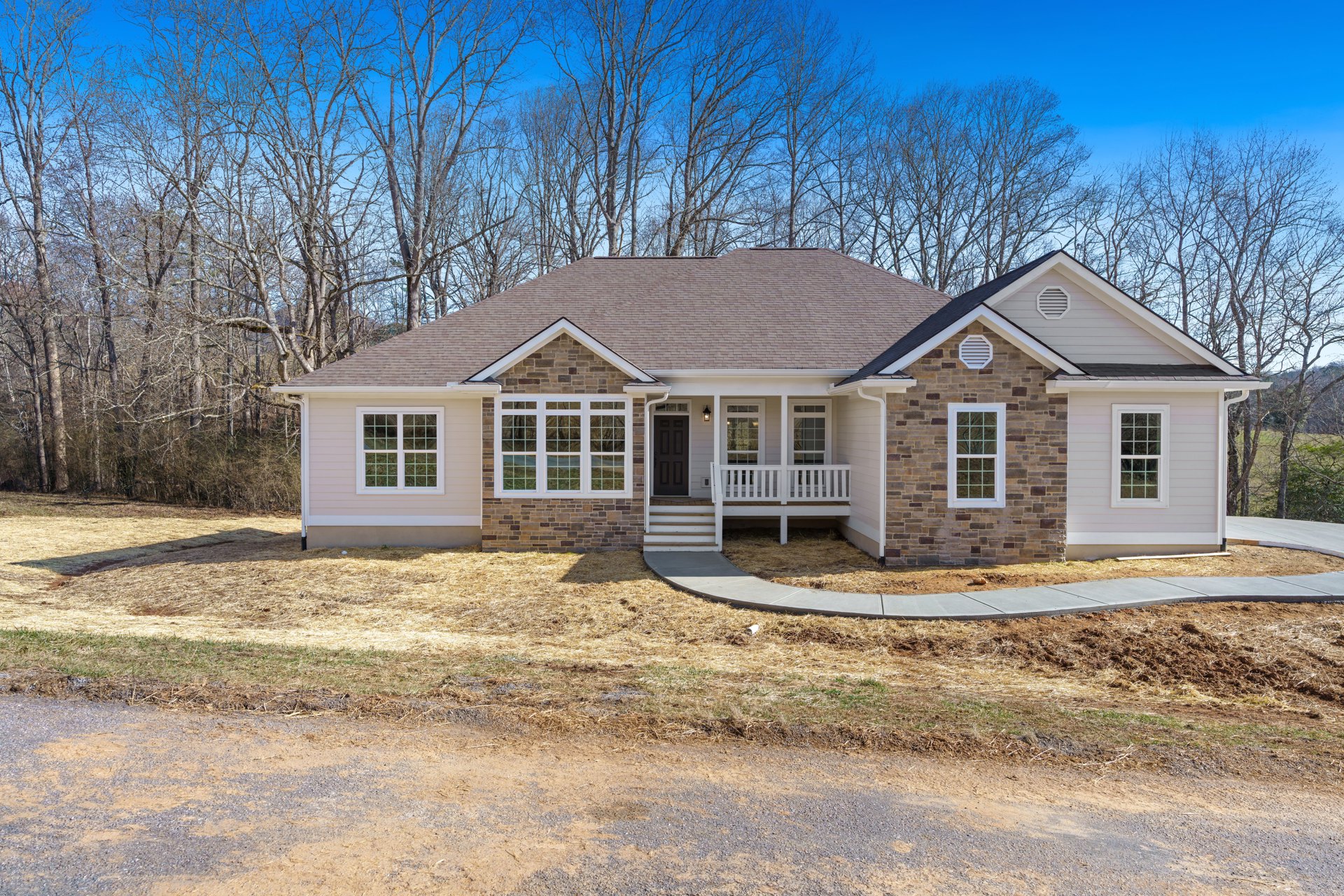 Two-story home with gray siding, white porch railing, large windows with white frames, paved driveway, mature trees in the background