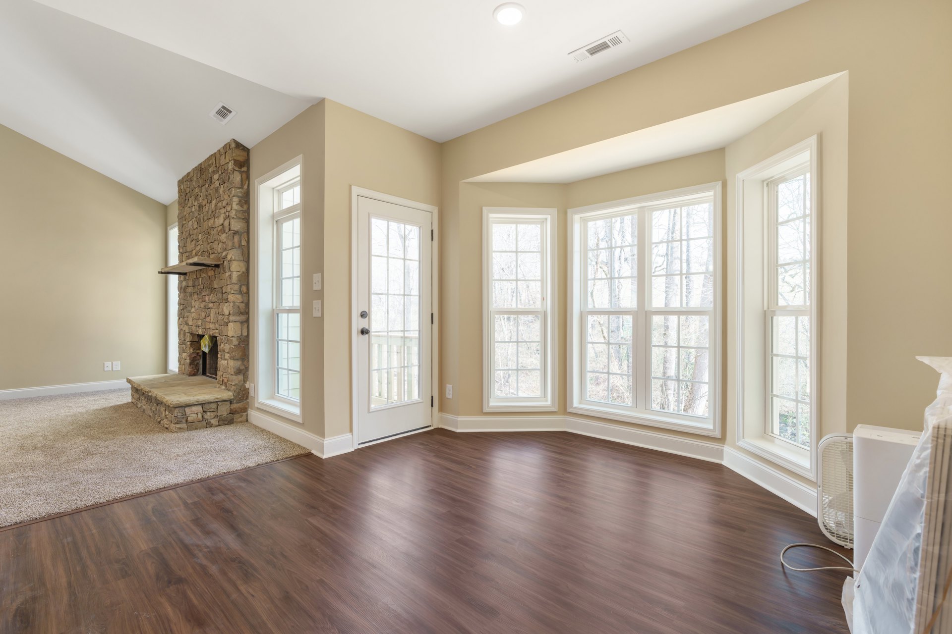Living room with hardwood floors, stone fireplace with stone ledge, multiple large windows showing tree views, and glass door with metal knob