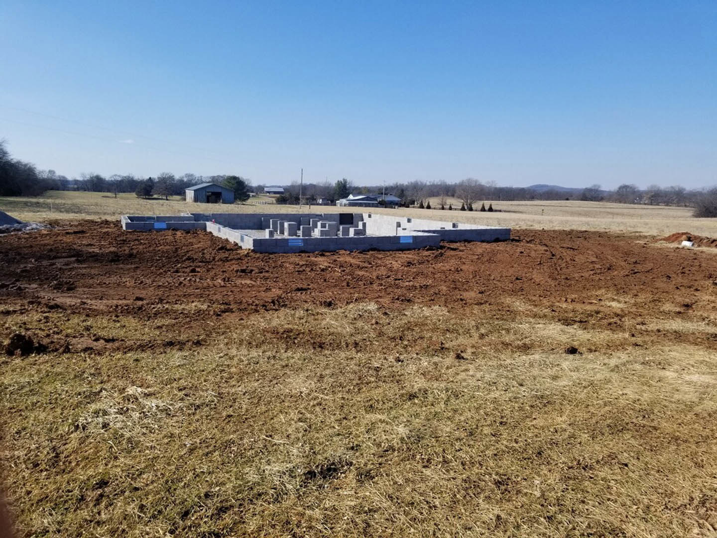 Concrete foundation and stacked blocks on a dirt field with grass, white can nearby, blue sky and clouds overhead, trees in the background