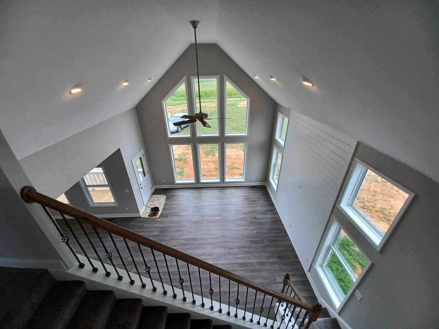 Wood staircase with white balusters and handrail, ceiling fan above, large windows letting in daylight, dog resting on hardwood floor, pole in open room, window overlooks dirt