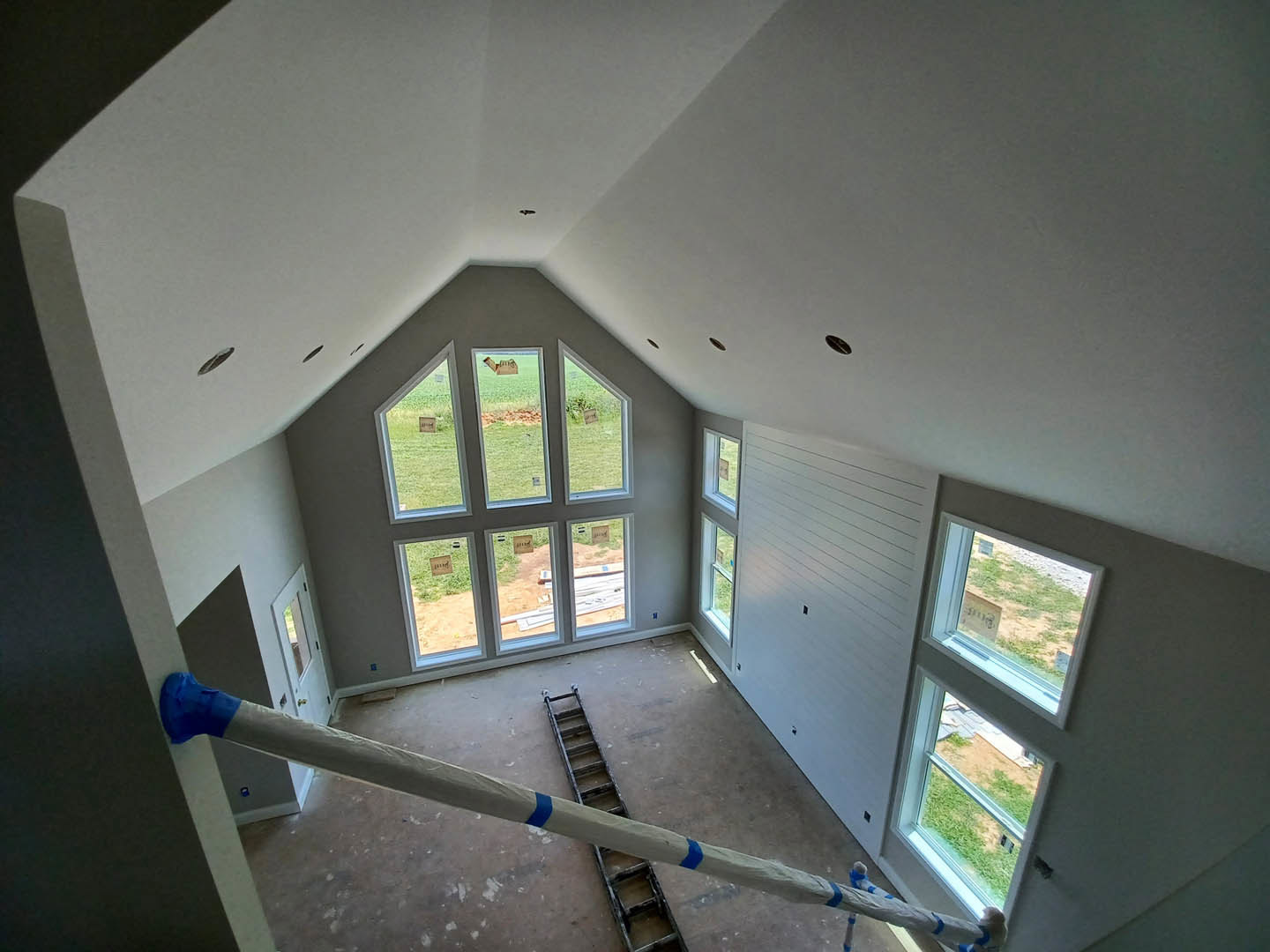 Bright room with white plaster walls, multiple tall windows with stickers, wooden ceiling, and several ladders positioned near a long pole; green field visible through the windows.