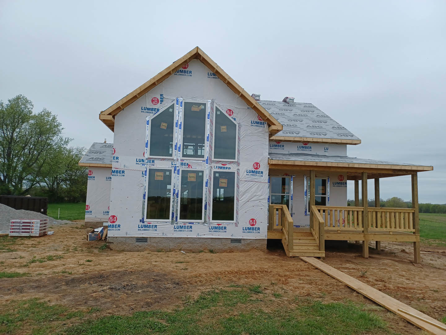 Partially built house with covered porch, exposed wooden framing, stacked bricks along exterior wall, wooden deck with railing, green trees in background, scattered lumber on