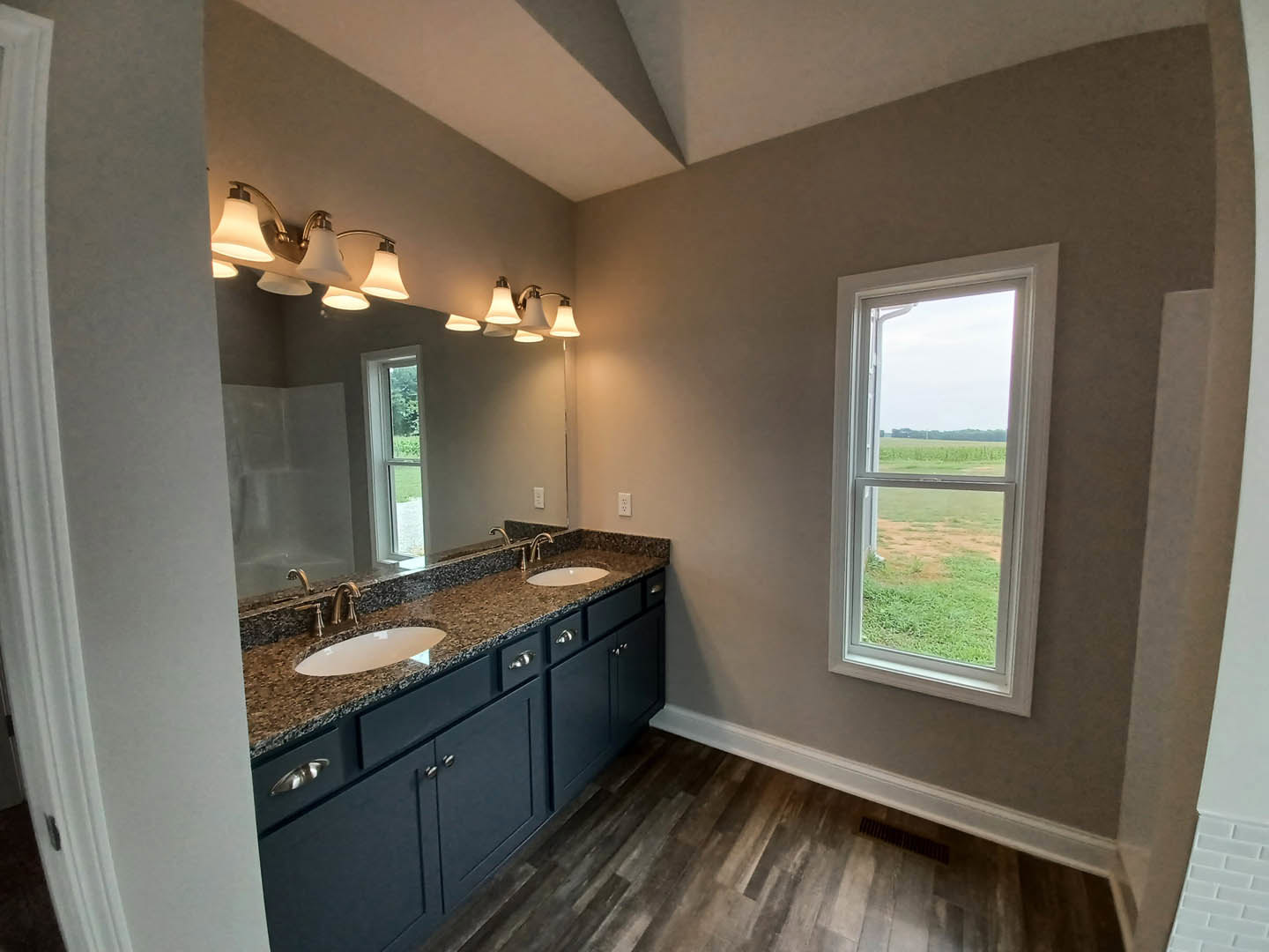 Bathroom with double sinks set in a marble countertop, large frameless mirror above, tile flooring, cabinetry below, and window overlooking grassy field.
