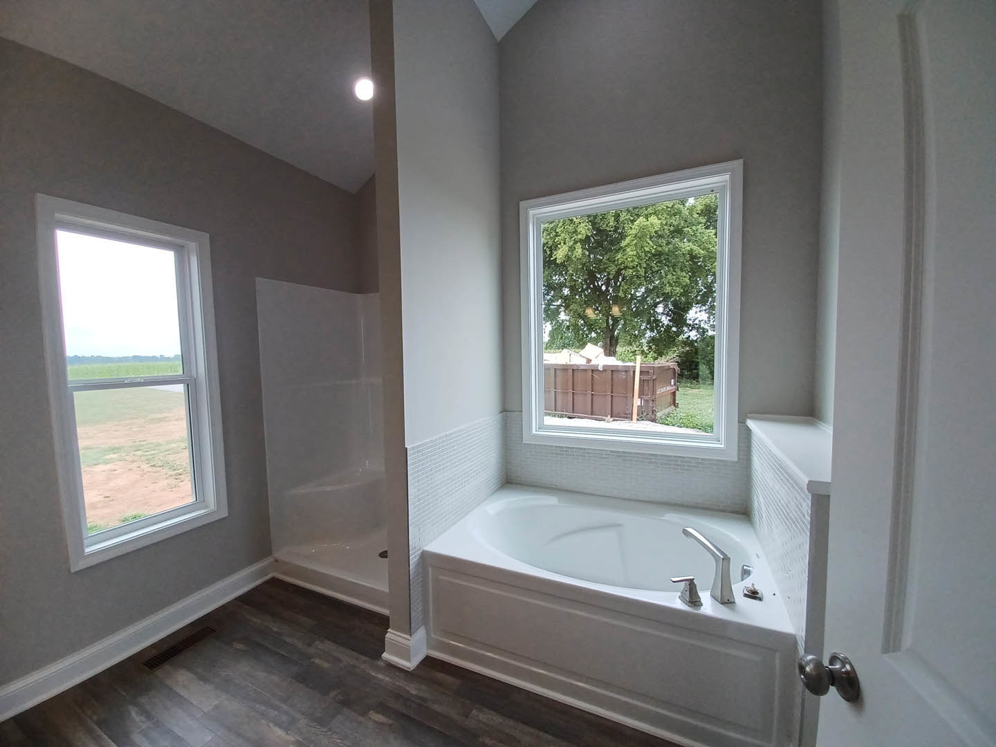 Freestanding white bathtub with chrome faucet beneath large window overlooking grassy field, light-colored walls, and brown storage bin on floor