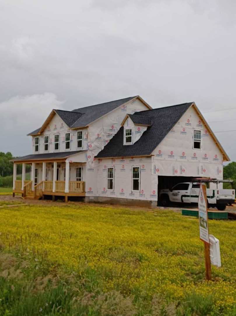 Partially built farmhouse with exposed wooden framing, broken roof, white truck parked behind, grassy lot with yellow wildflowers, cloudy sky overhead