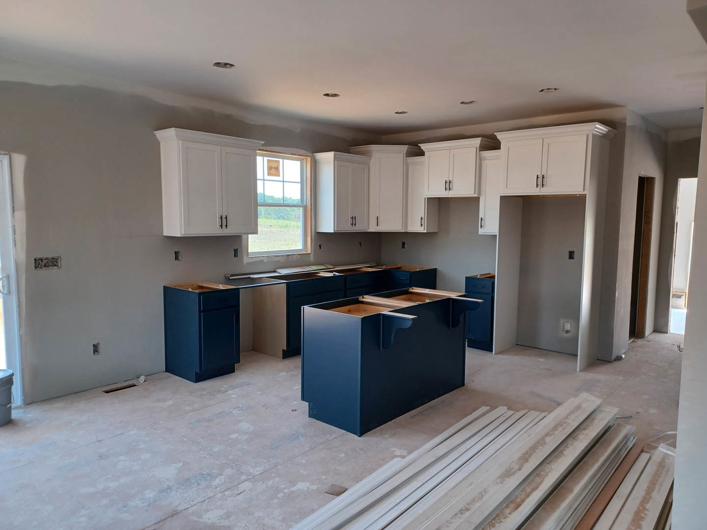 White kitchen cabinets with black handles, blue countertops, blue lower cabinet with drawer, two-shelf blue rectangular island, stainless steel sink, window with sign, pile of