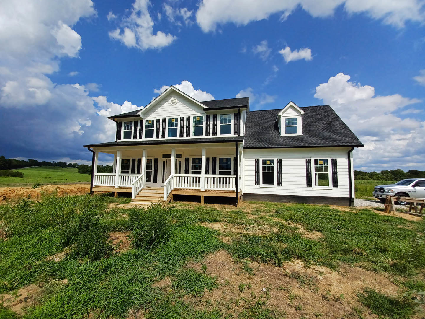 White two-story house with black shutters, expansive covered porch, silver car parked on driveway, manicured grassy lawn, blue sky with scattered clouds