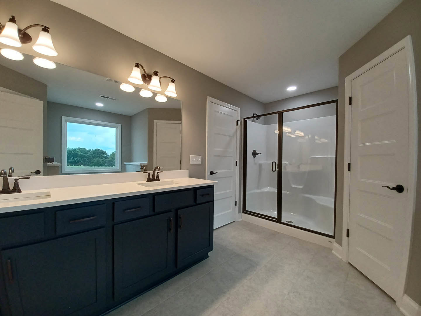 Bathroom featuring a glass shower enclosure, white sink with chrome faucet, wall-mounted mirror, white door with black handle, and window overlooking trees.