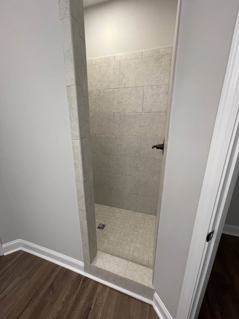 Glass shower enclosure with black handle, white tile walls, wood flooring outside shower, tile shower floor with central drain