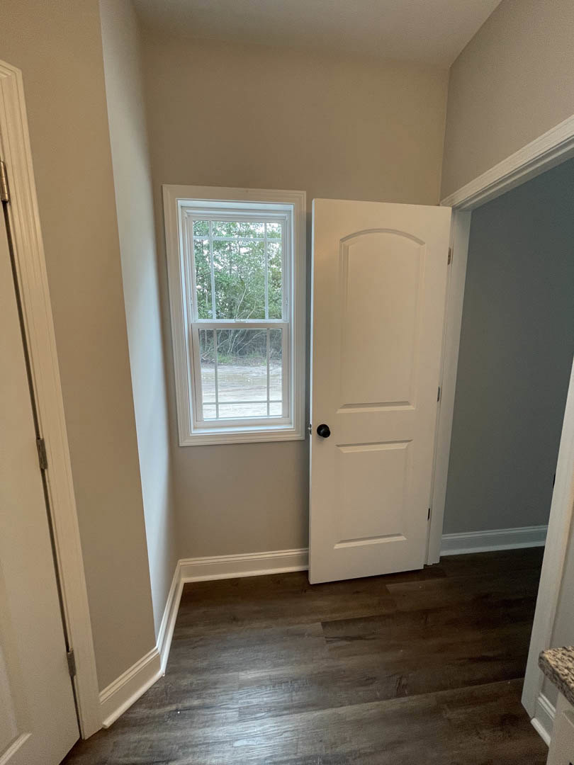 White paneled door with brushed metal handle, dark wood flooring, white baseboard trim, adjacent window framing leafy trees outside