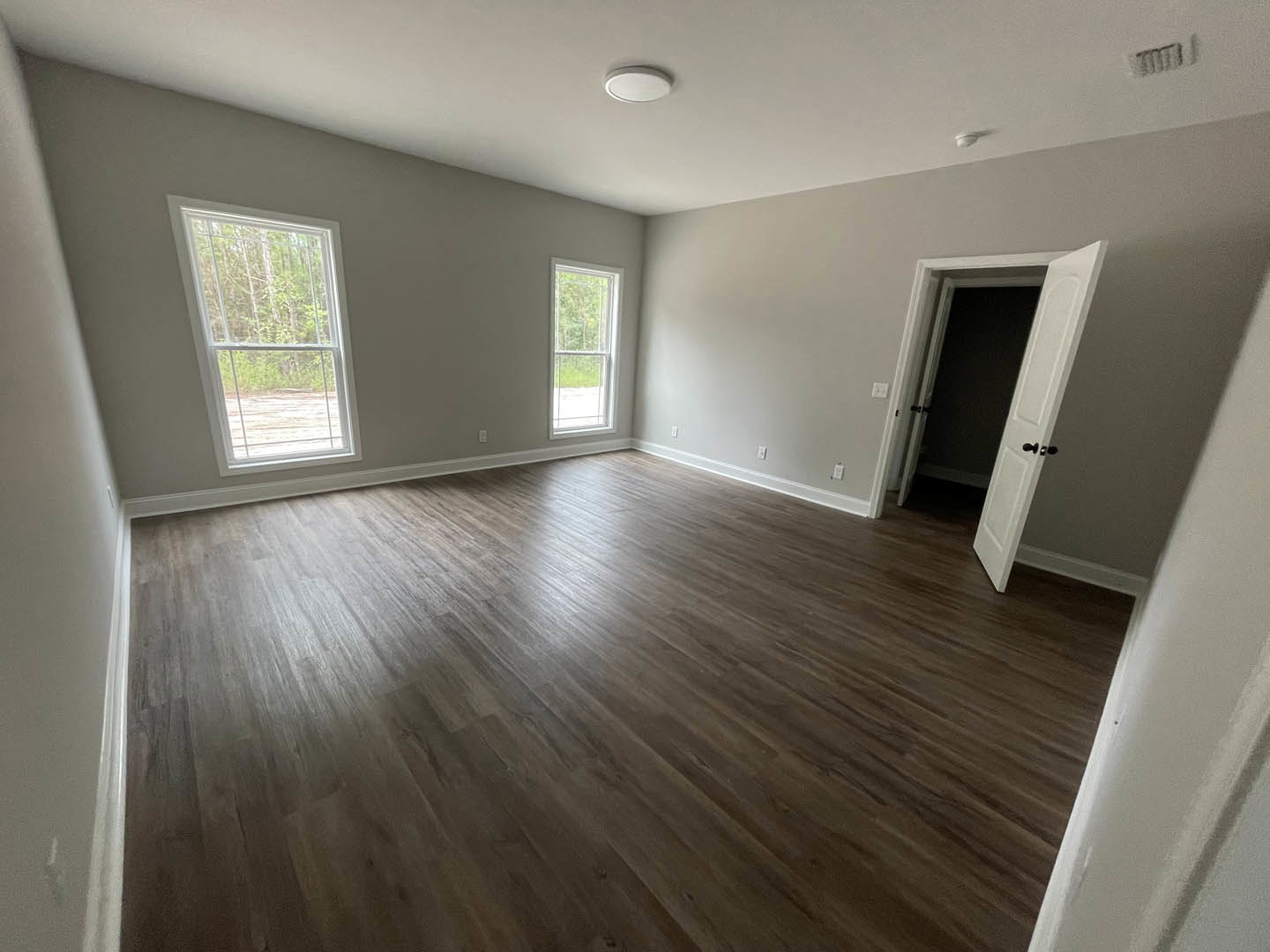 Room with light wood flooring, white plaster walls, large window with white frame showing trees outside, and a single door; white circular fixture on wall and numbered plaque