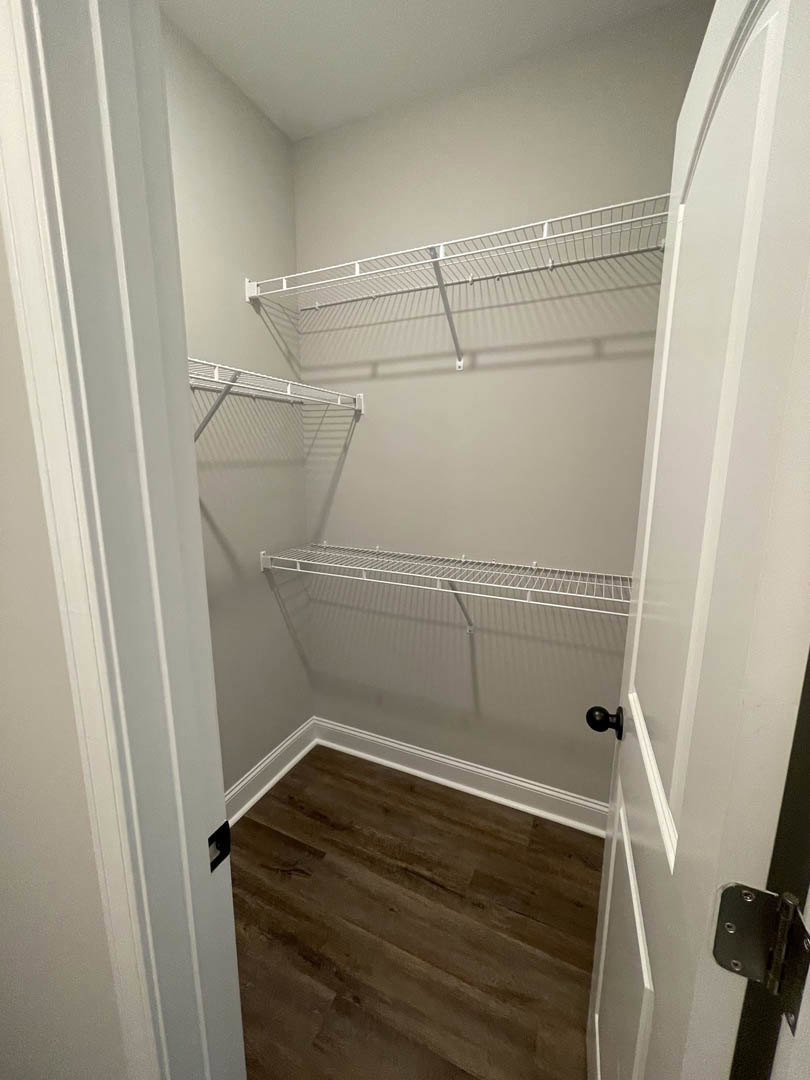 Walk-in closet with white shelving, dark wood flooring, white trim, and a white door with metal shelf supports and visible hinge