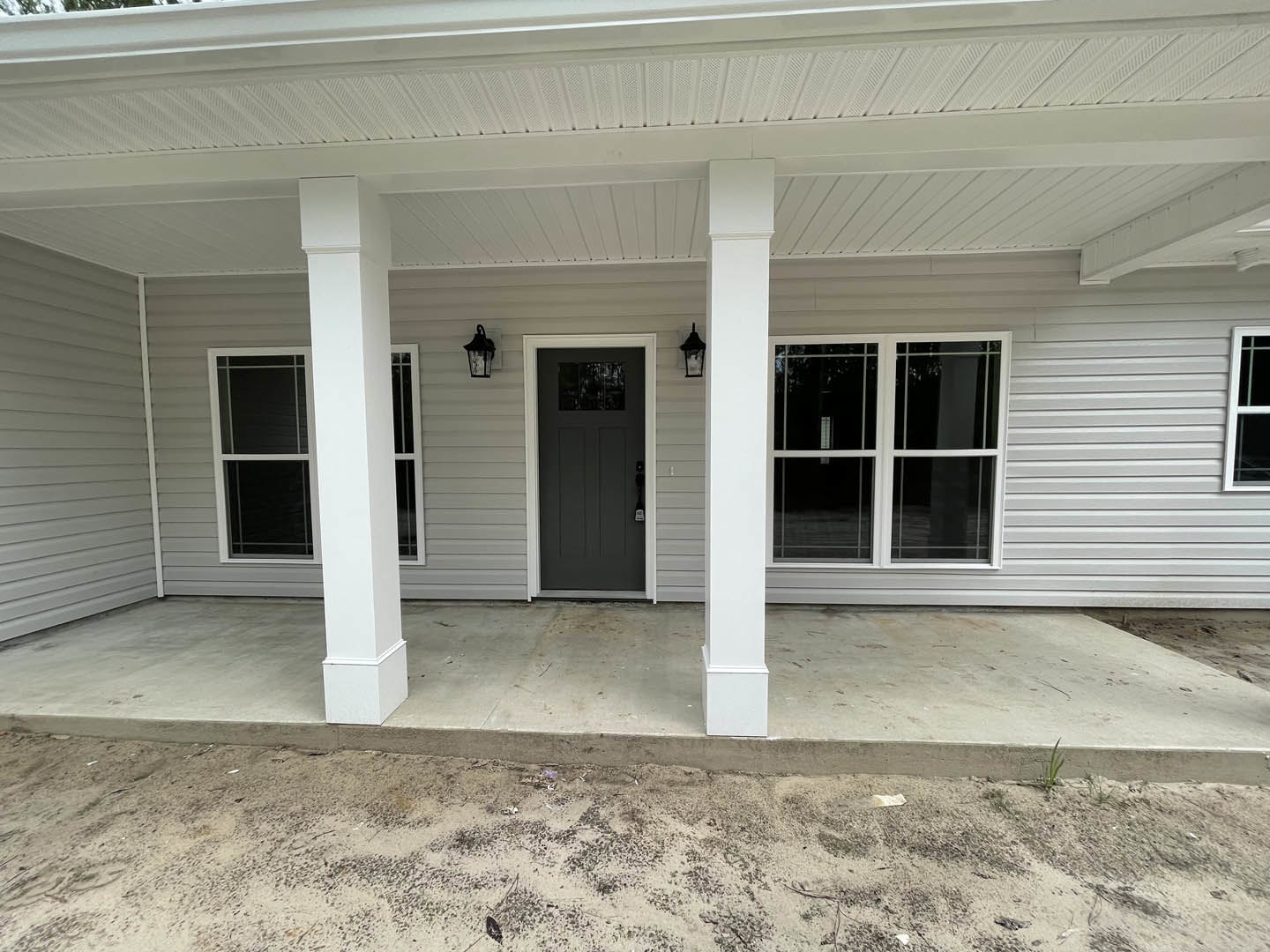 Modern home exterior with light siding, two grey entry doors, white-framed windows, covered porch ceiling, and dirt ground in front