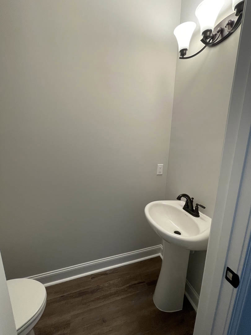 White pedestal sink with black faucet beside a modern toilet, light fixture above, gray tile walls, and rectangular mirror reflecting bathroom finishes.