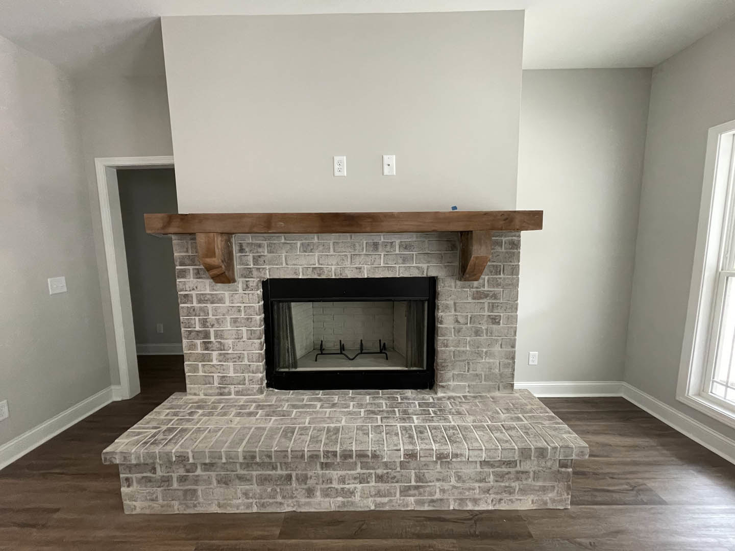 Black-framed fireplace set into a brick accent wall with wood mantel, white walls, window, and dark fire screen on light flooring